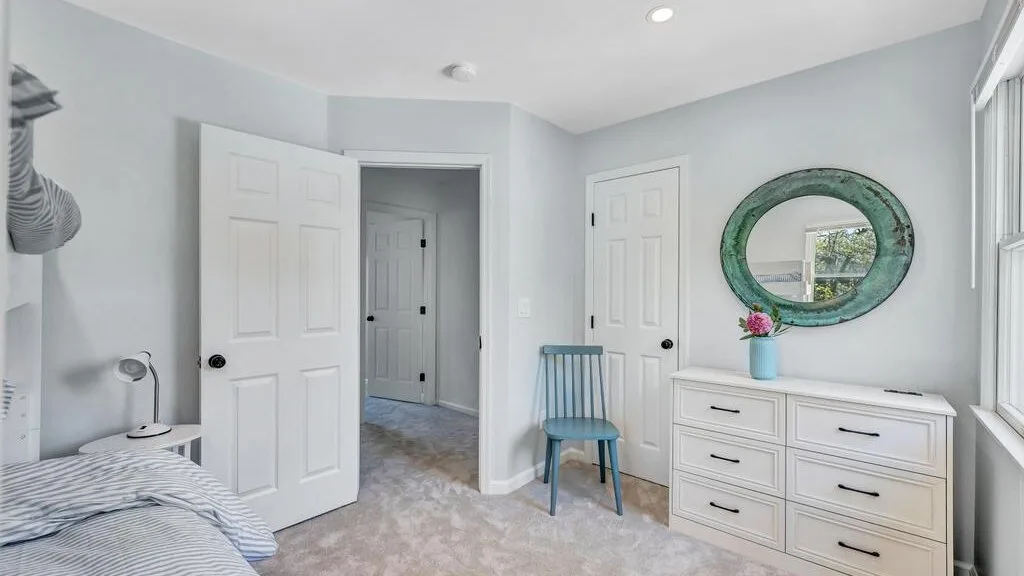 A bright bedroom with a bed, a blue chair, and a dresser beneath an oval mirror.