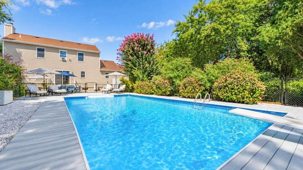 An outdoor swimming pool bordered by a deck, shrubs, and a house.