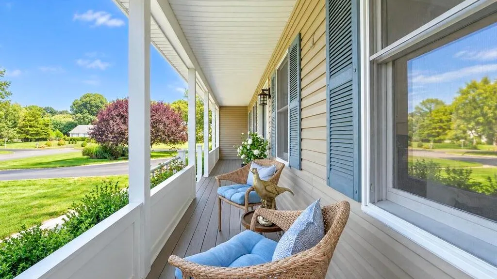 A covered porch with cushioned seating overlooks a green lawn and trees.