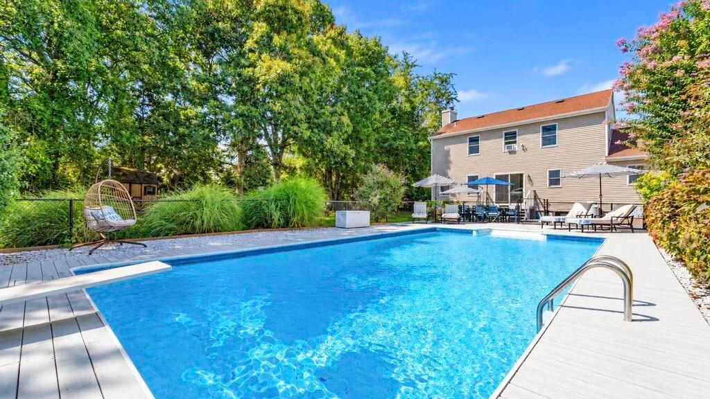 An outdoor swimming pool sits beside a deck, with a house and trees in the background.