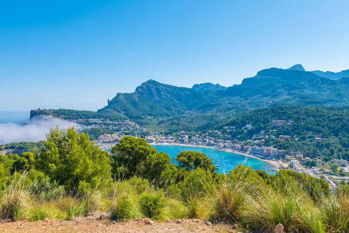 Una bahía costera y un pueblo se encuentran bajo montañas verdes, vistos desde una ladera.