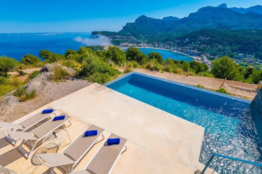 Una piscina al aire libre y tumbonas miran a una bahía costera y una costa montañosa.