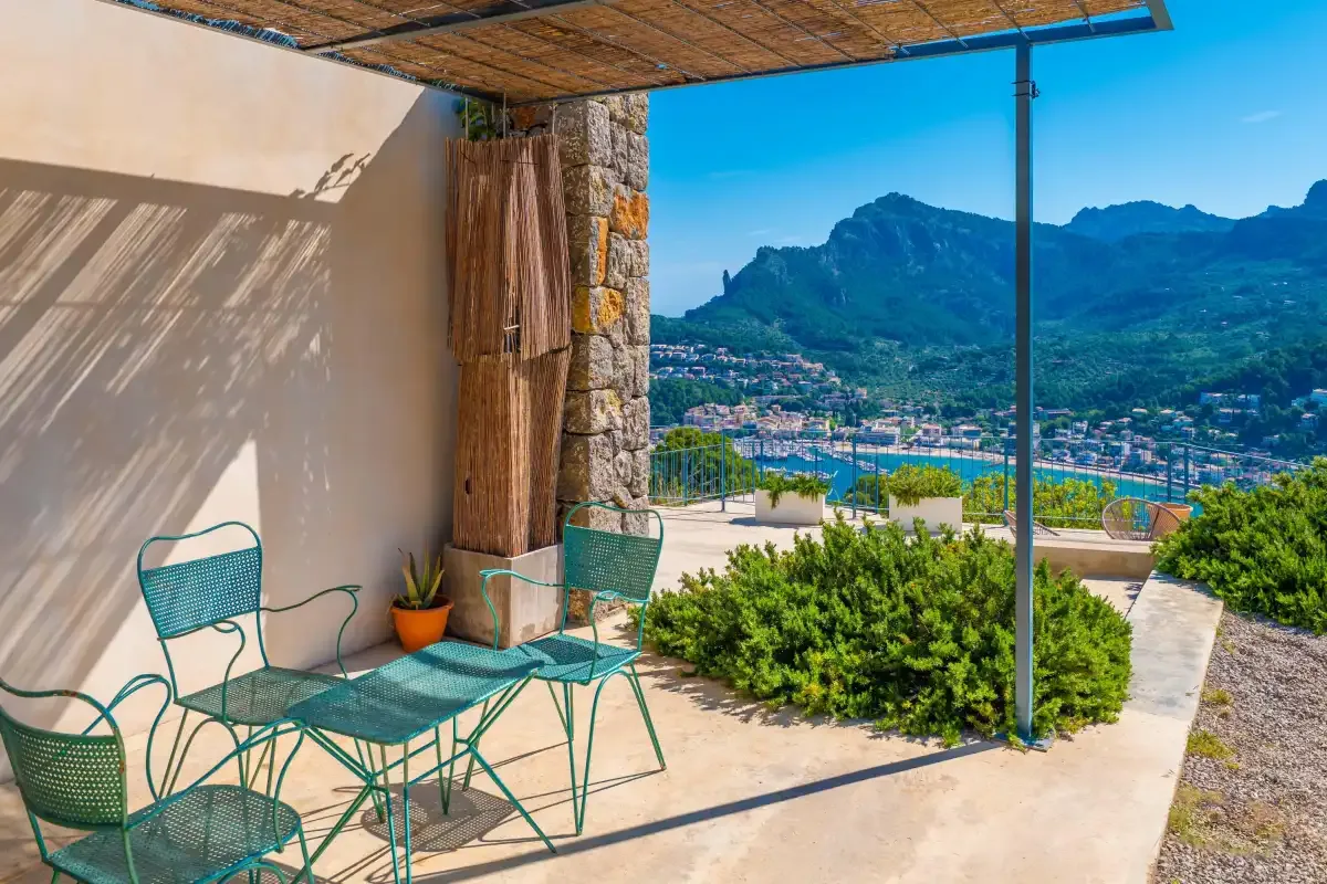 Una terraza sombreada con asientos metálicos ofrece vistas a un pueblo costero y montañas.