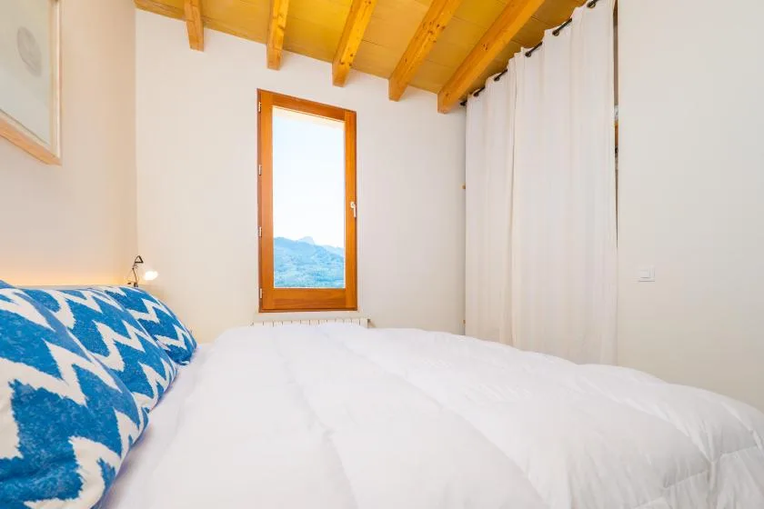 Un dormitorio luminoso con ropa de cama blanca, un cojín estampado azul y una ventana bajo un techo de vigas de madera.