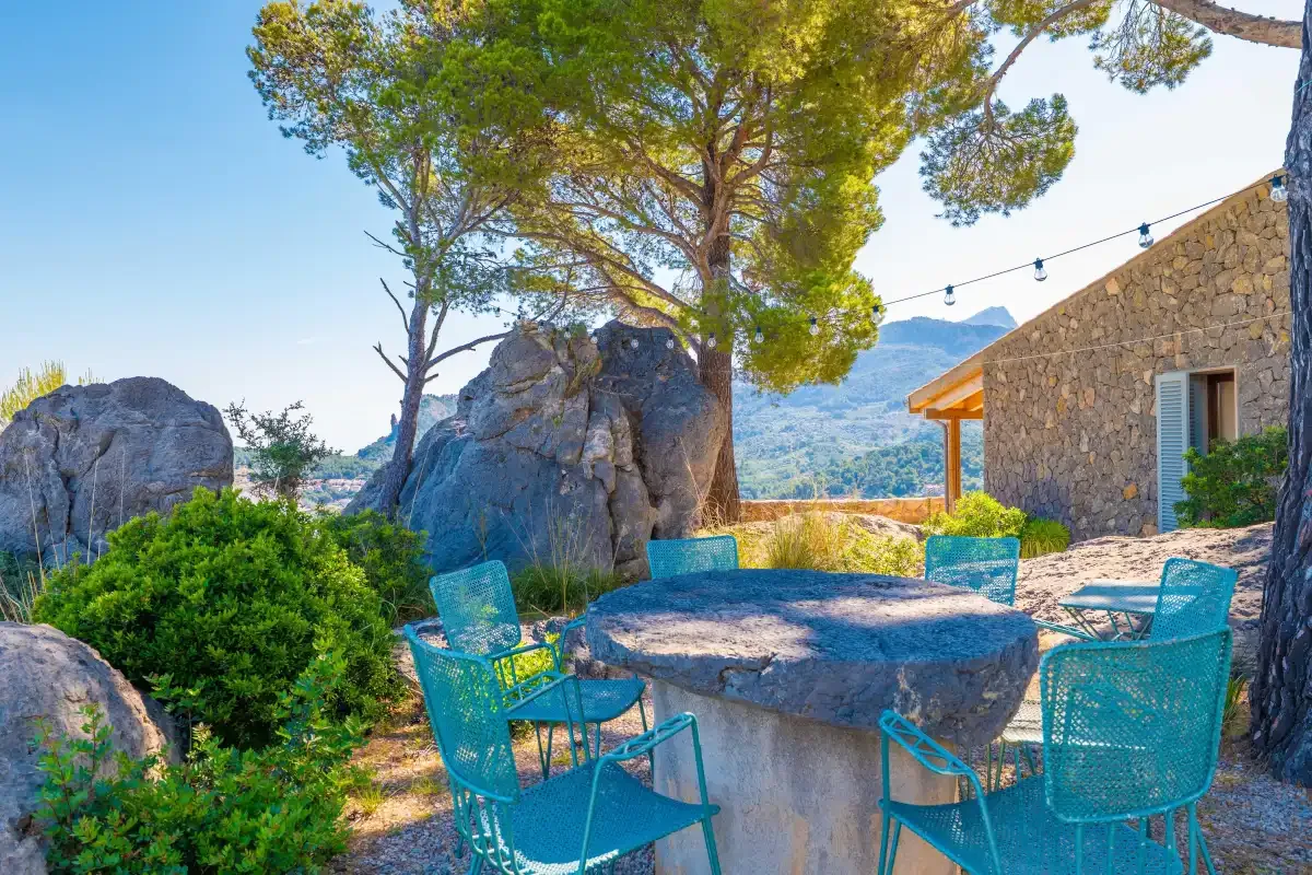Un patio al aire libre con asientos turquesa rodea una mesa de piedra bajo un árbol grande.