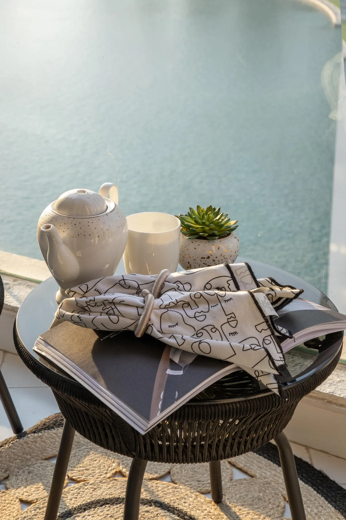 A small round table holds a teapot, folded fabric, and a potted succulent beside a pool.