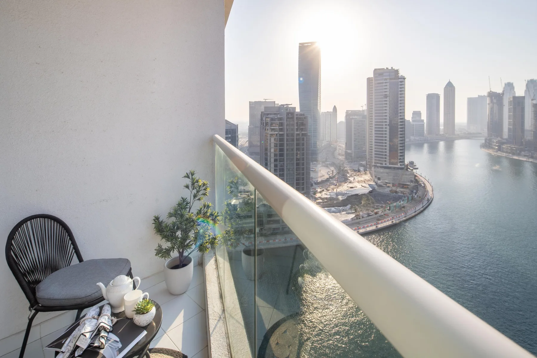 A balcony seating area overlooks a waterfront skyline, with potted plants beside the glass railing.