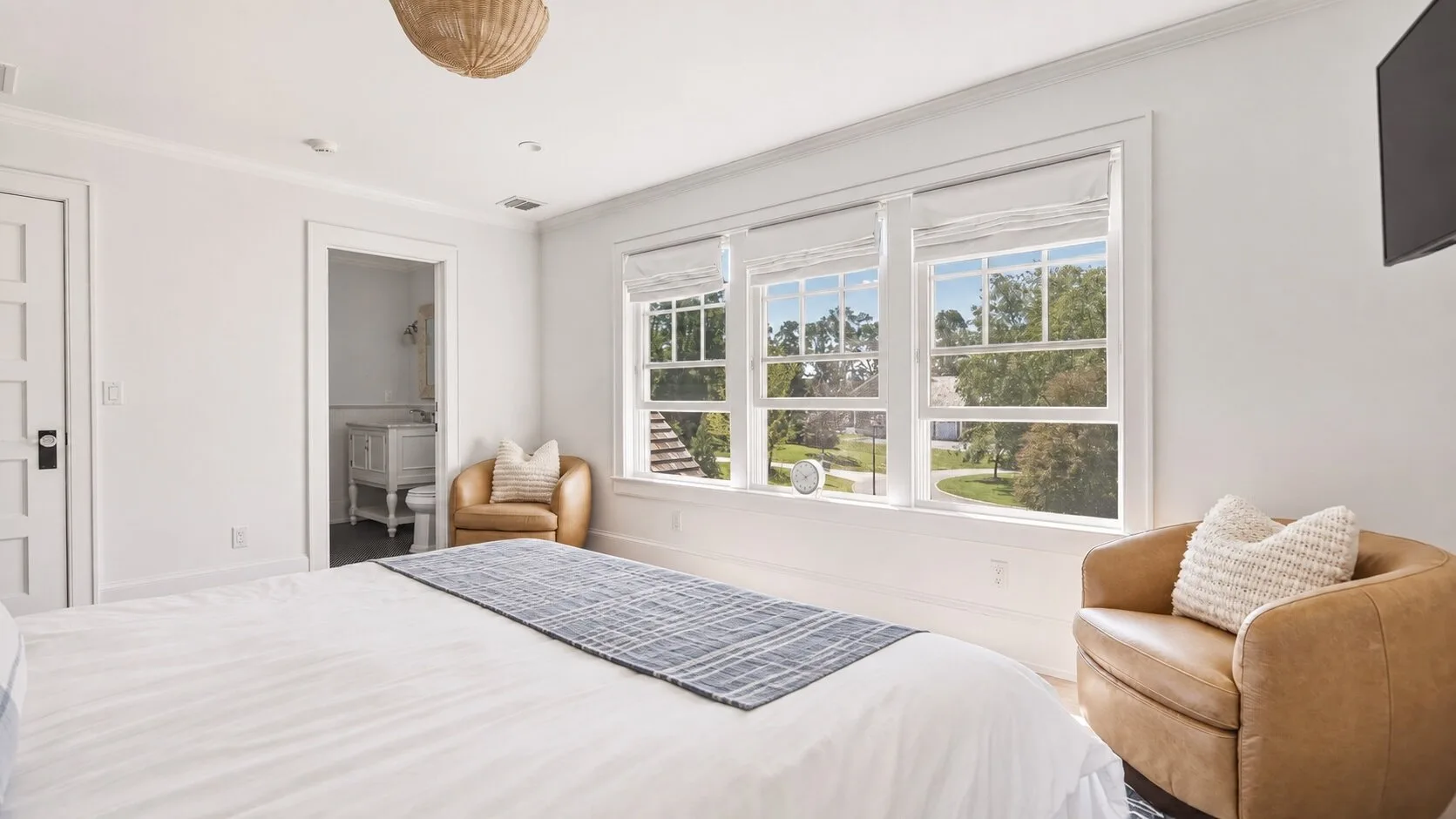 A bright bedroom with a large bed, a striped rug, and windows overlooking greenery.