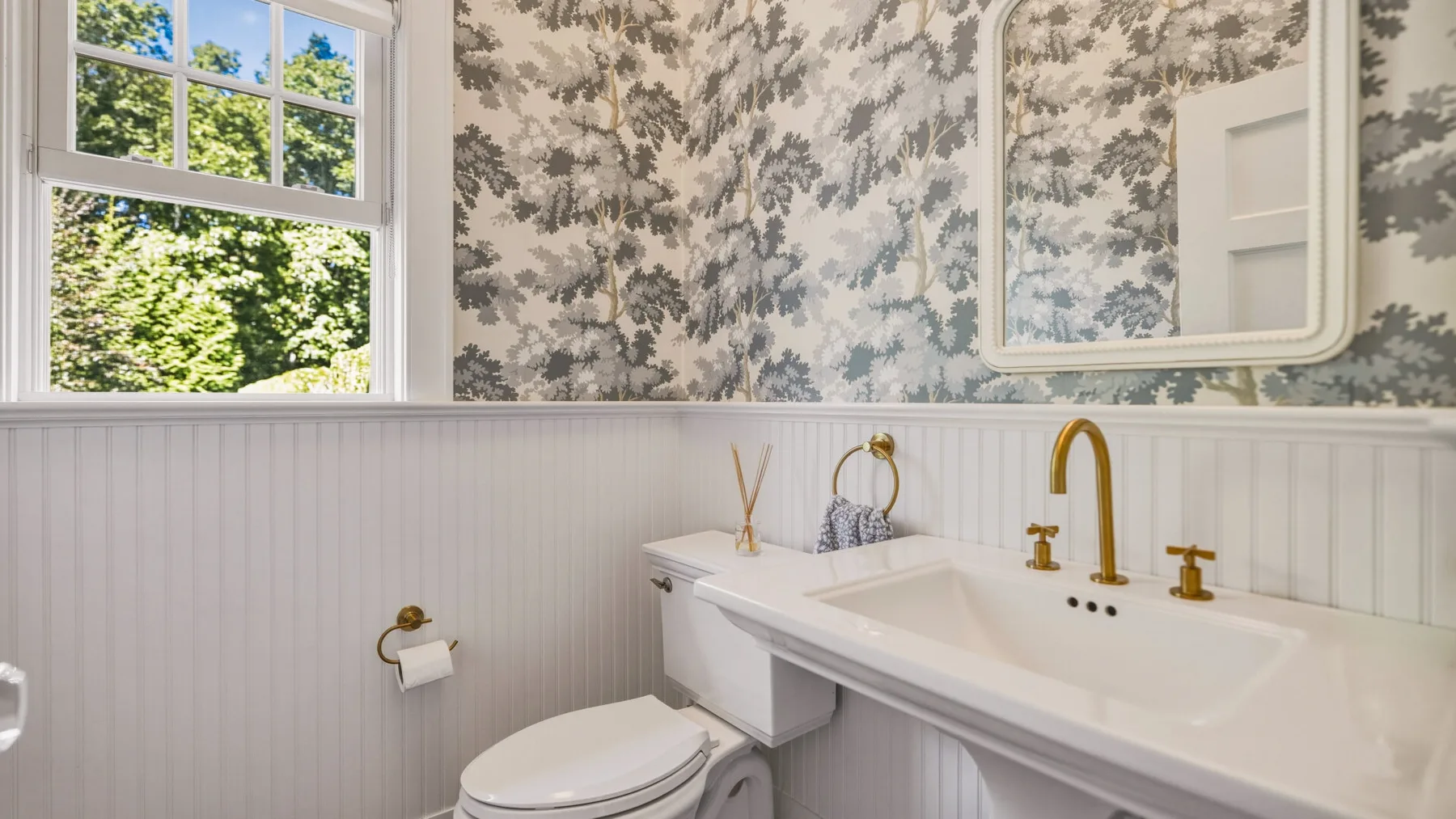 A bathroom with a long white sink beneath a mirror and floral wallpaper.