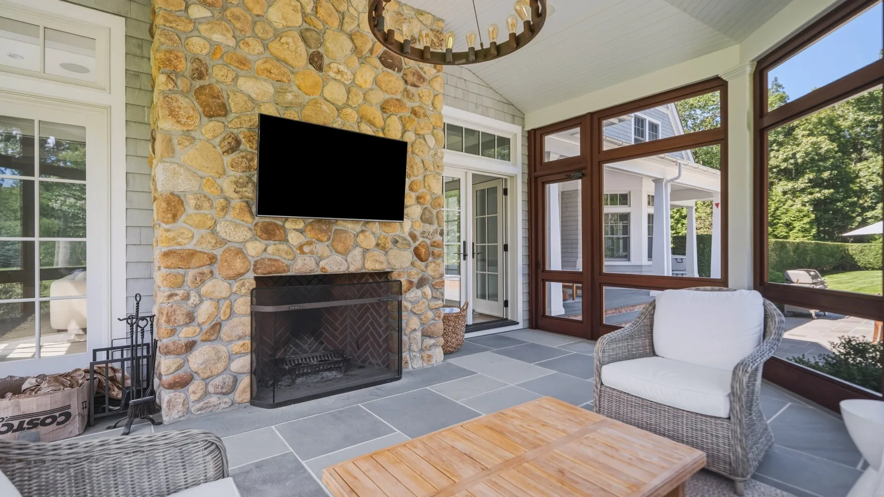 A bright sunroom has a stone fireplace, large windows, and wicker seating.