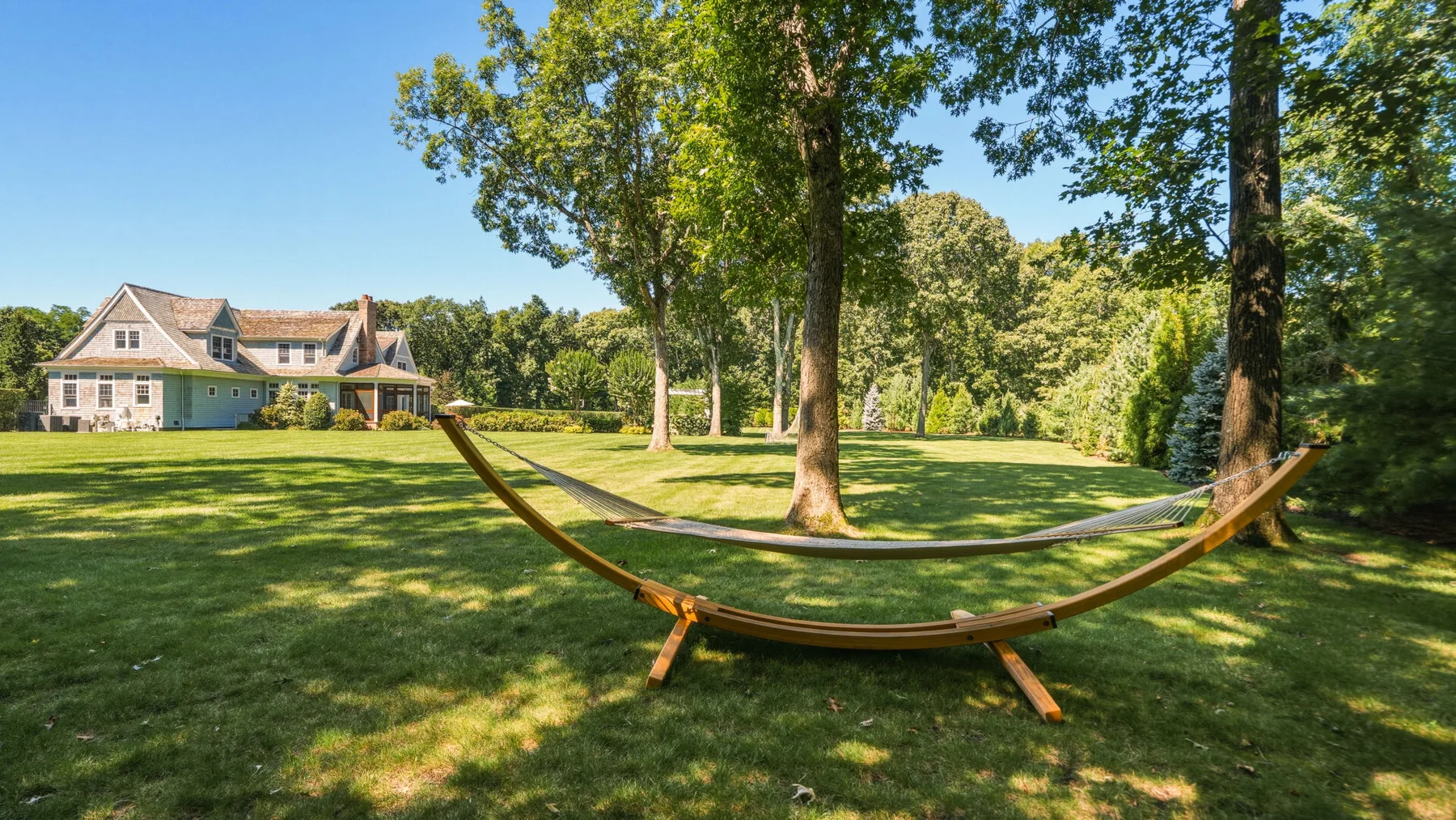 A hammock hangs between trees on a sunny lawn beside a house.