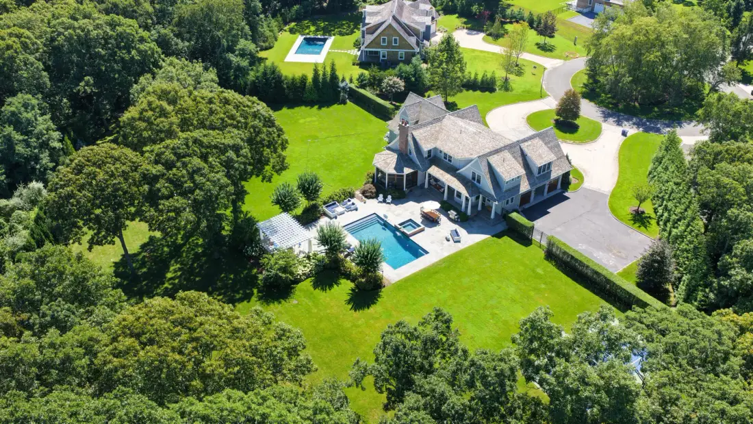 Aerial view of a large house with a pool, driveway, and surrounding greenery.