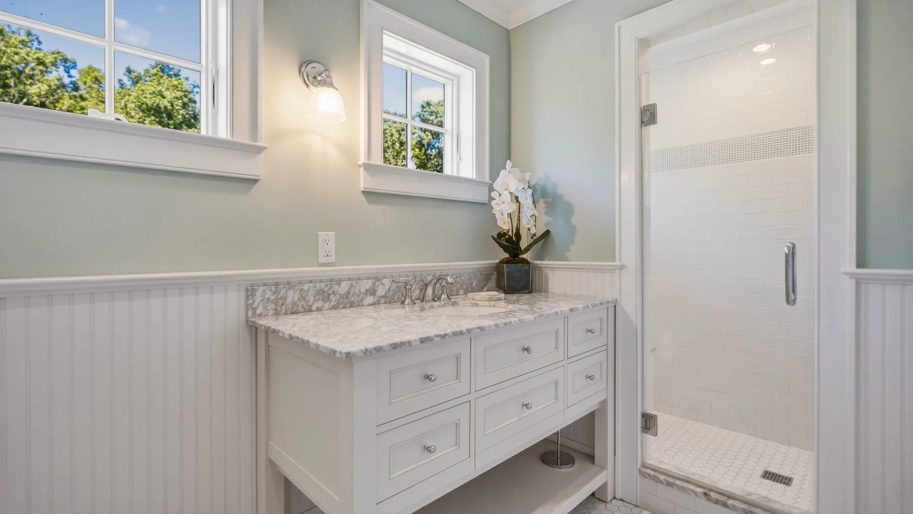 A bright bathroom features a white vanity beneath two windows beside a glass shower.