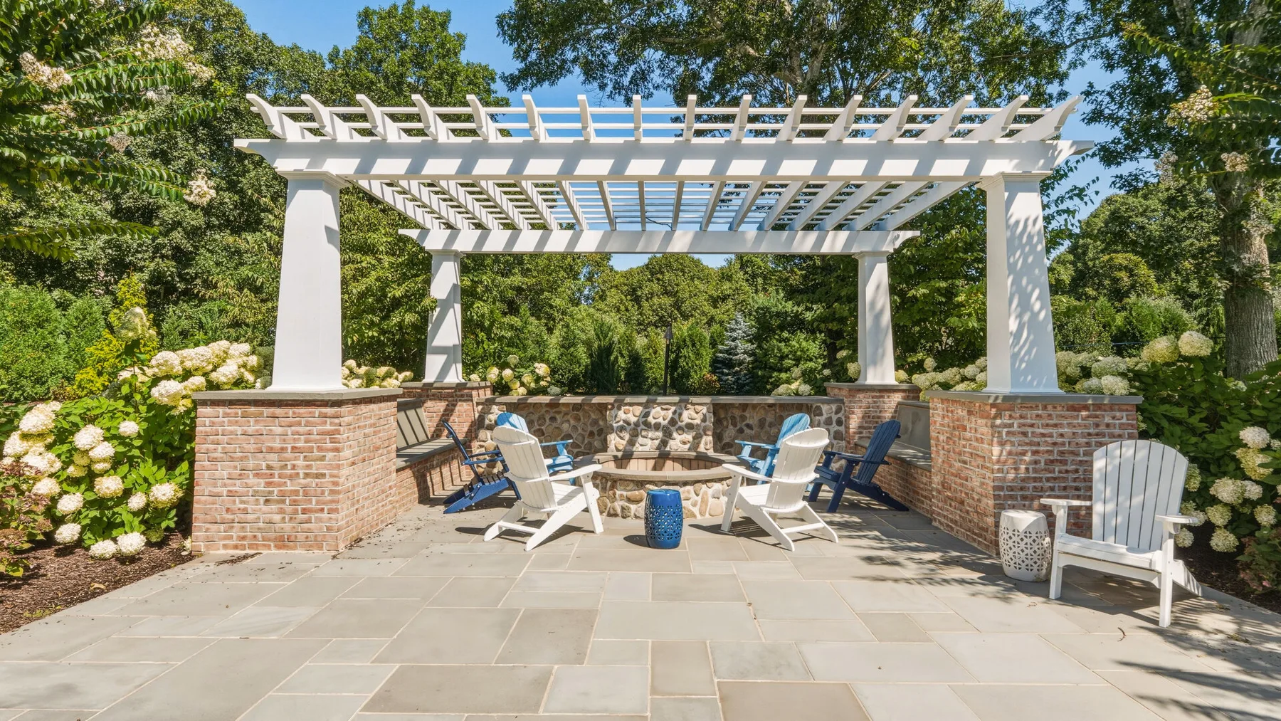 An outdoor patio with seating beneath a white pergola, surrounded by greenery.