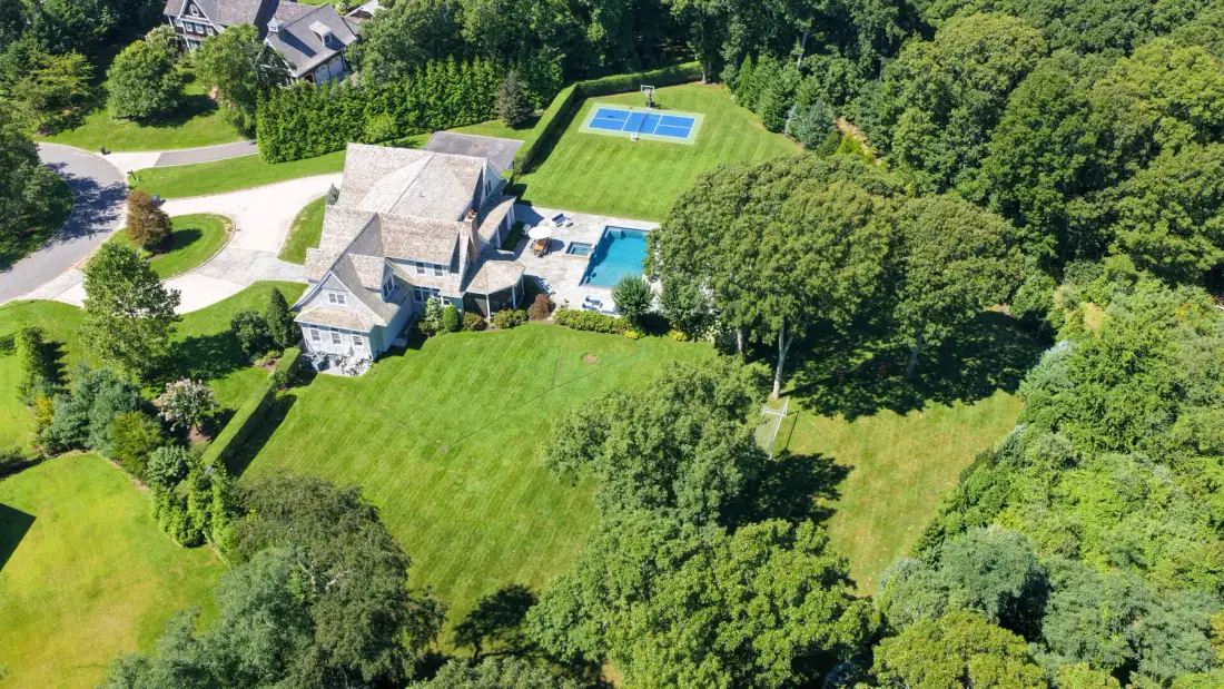 Aerial view of a house with a swimming pool and tennis court amid green lawns and trees.