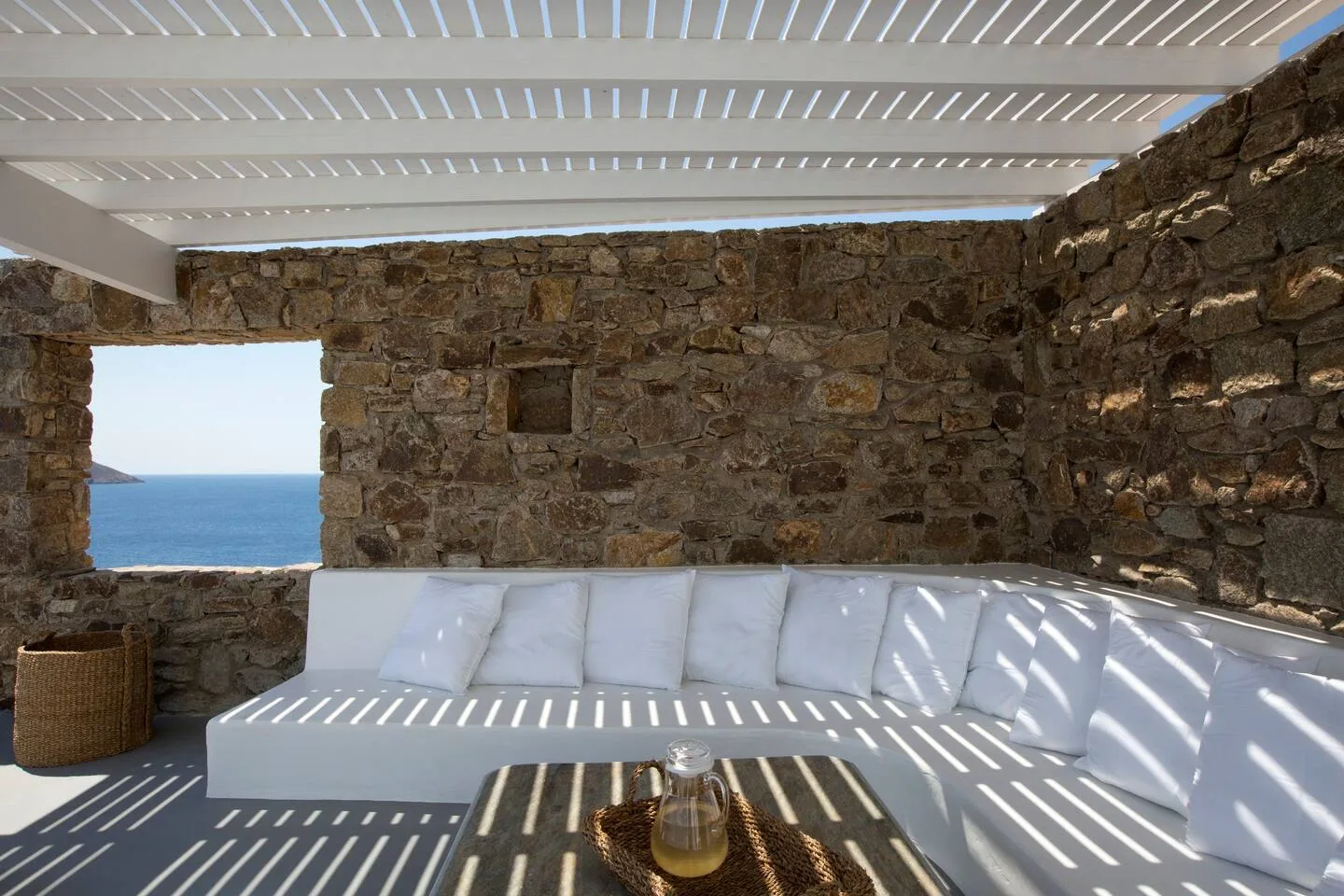 A covered stone patio has white seating and a view of the sea.