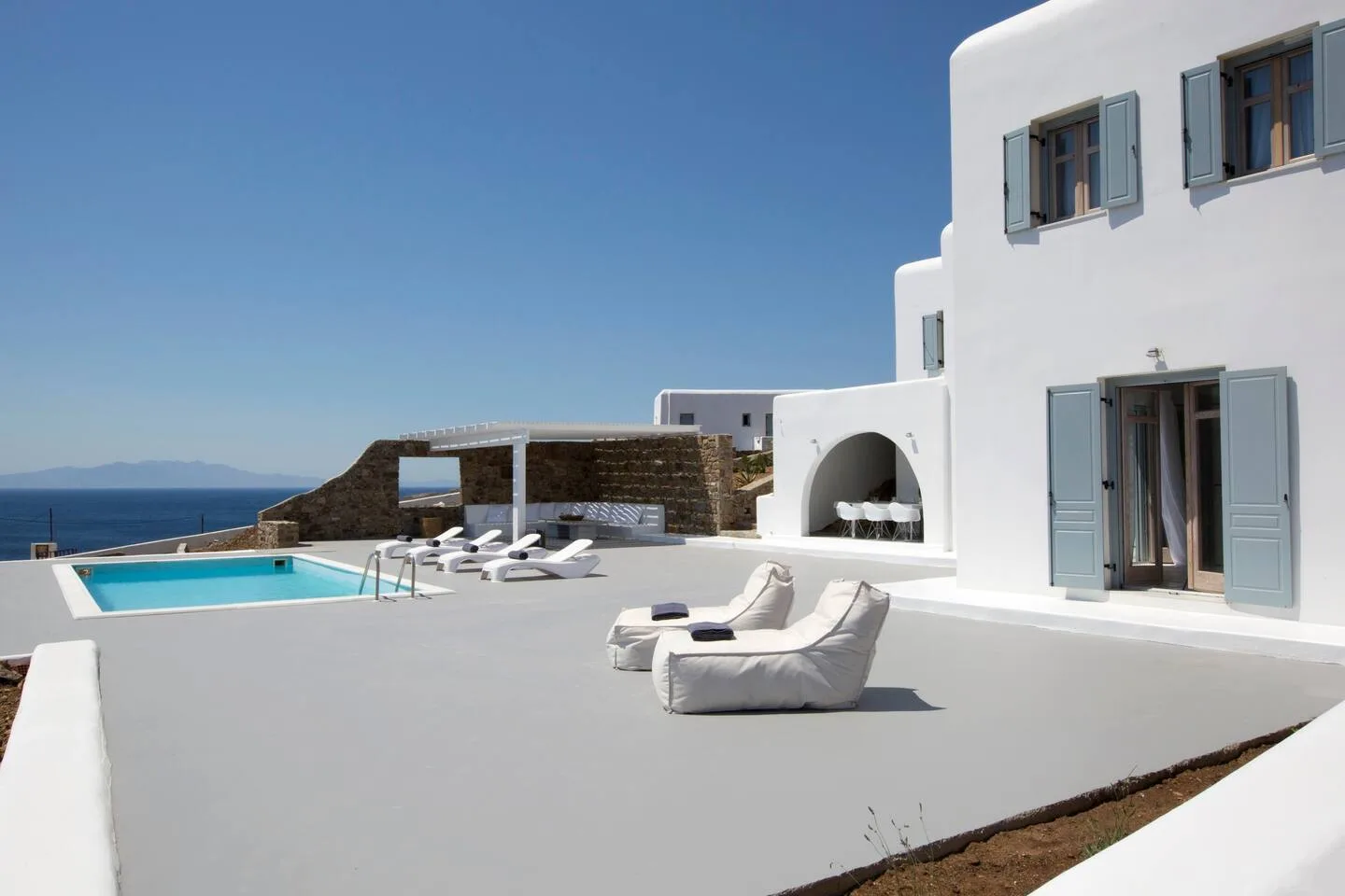 A white villa terrace features a pool, lounge seating, and a sea view.