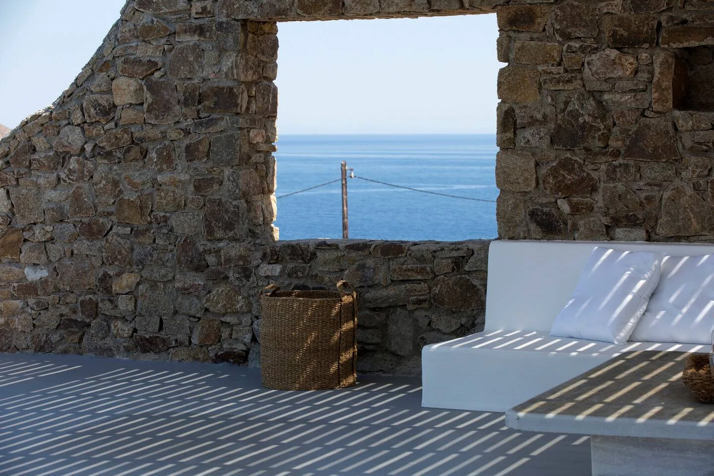 A stone terrace with white seating overlooks the sea through an open window.