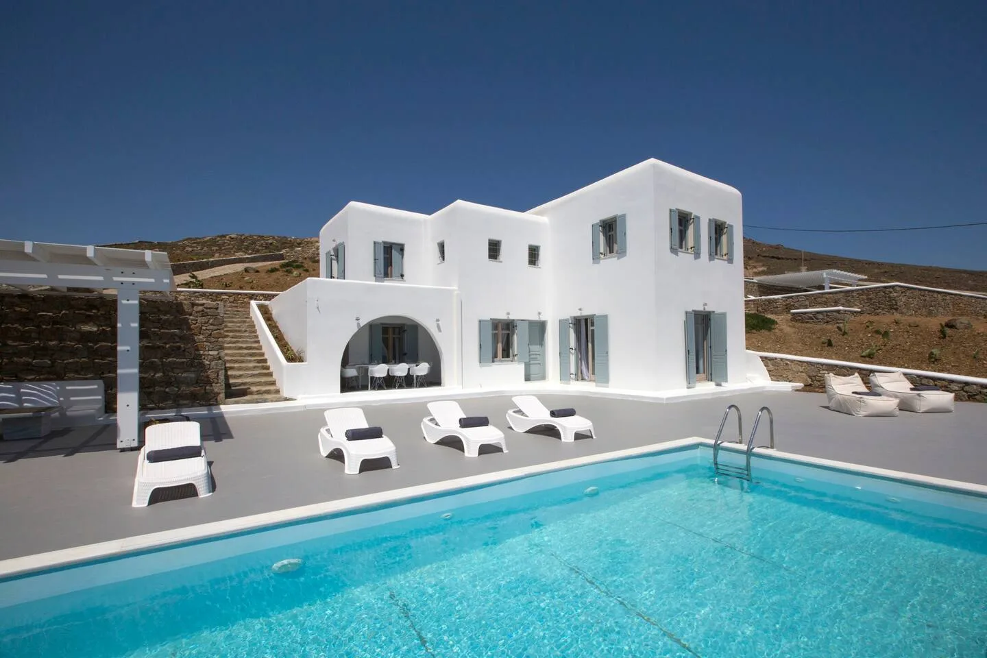 A white villa stands beside a swimming pool with loungers on the patio.