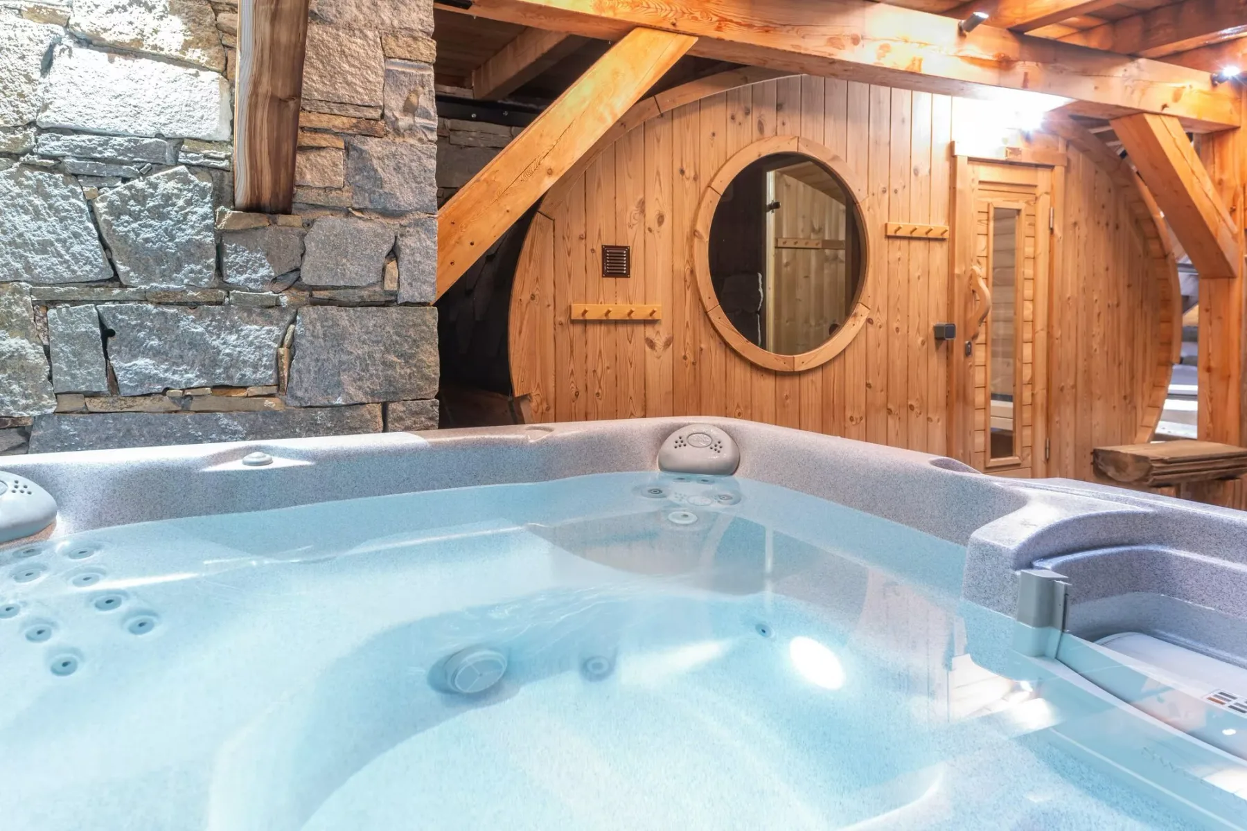 A hot tub sits in front of a wooden room with a round window and stone wall.