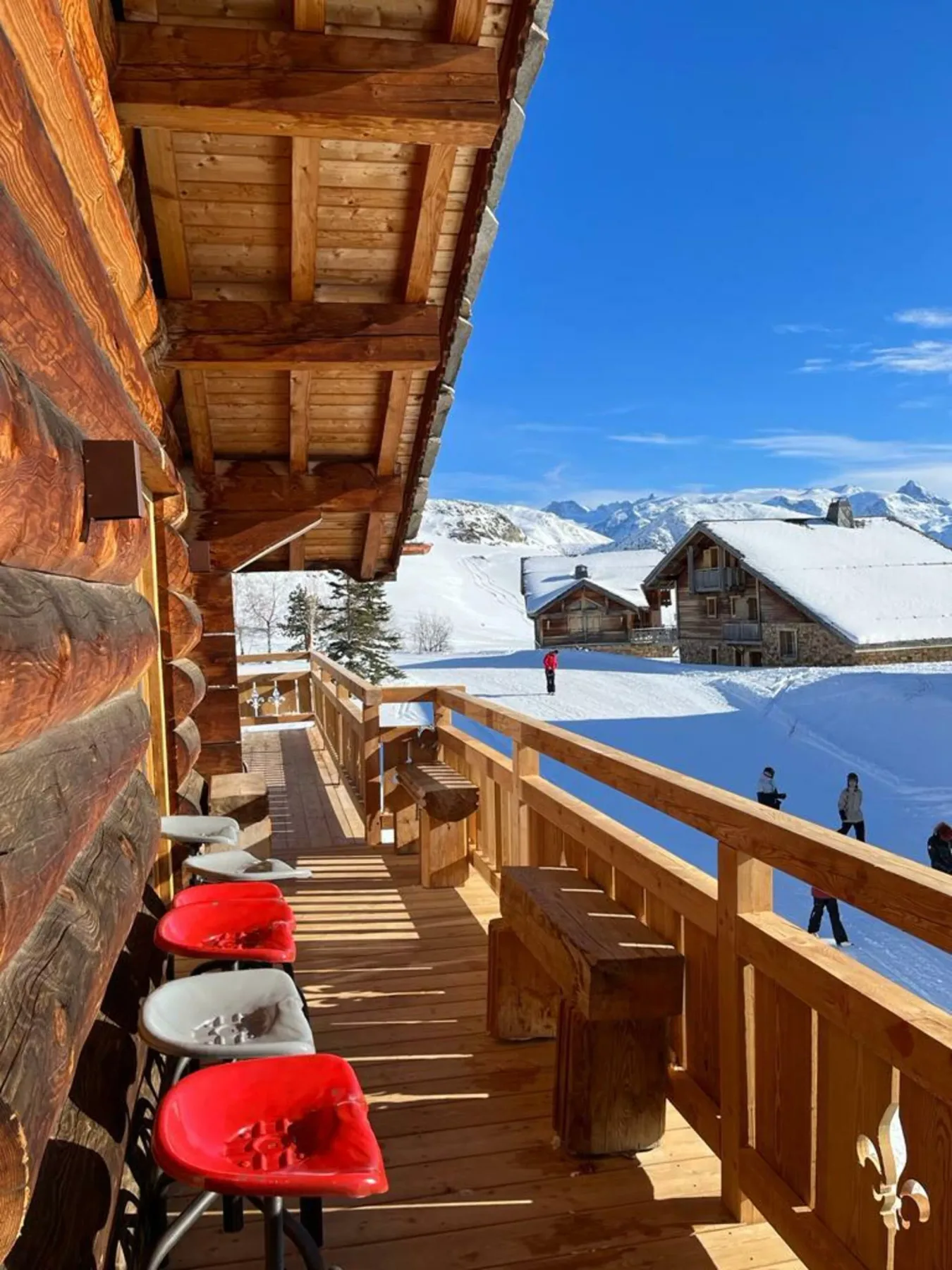 A wooden balcony overlooks snowy chalets and mountain peaks under a clear blue sky.