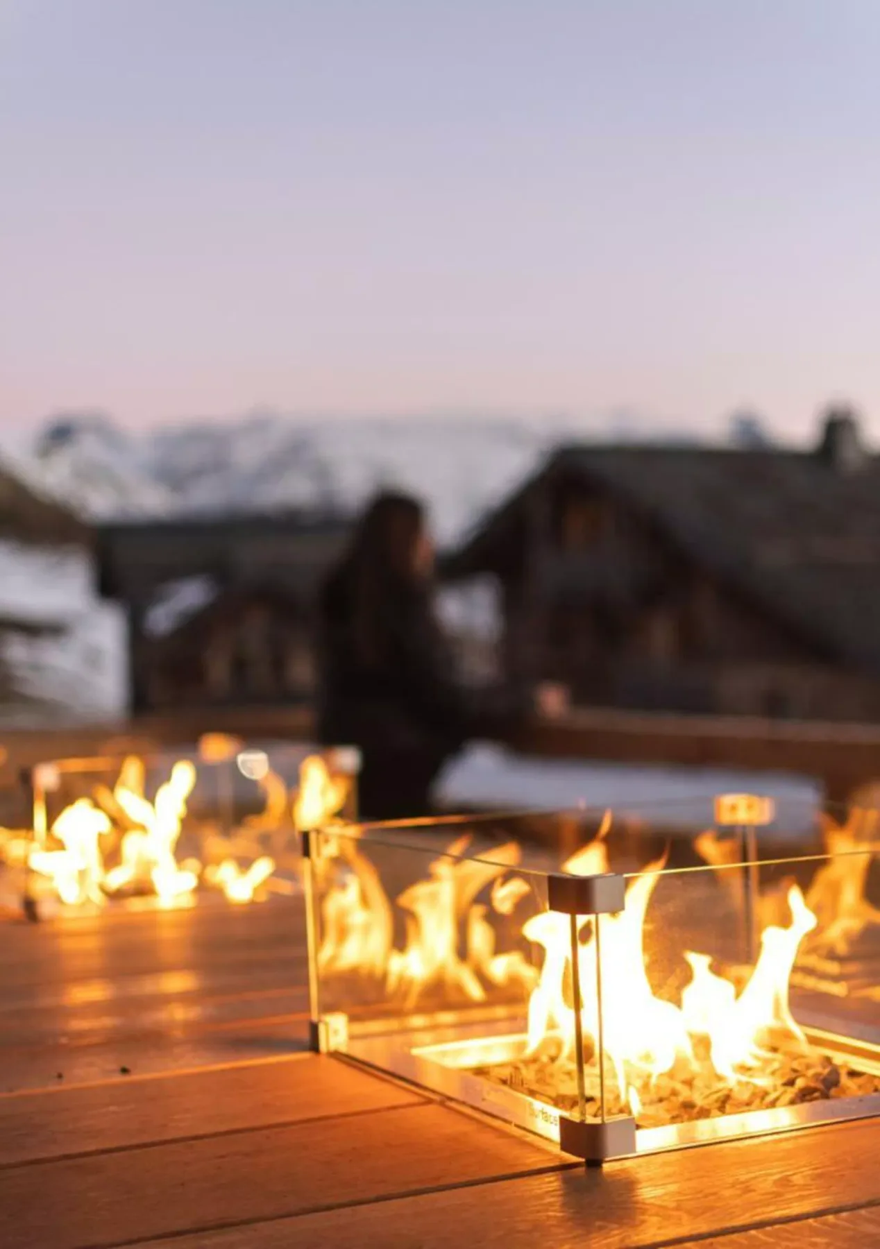 An outdoor fire feature glows on a wooden deck, with snowy mountains in the background.