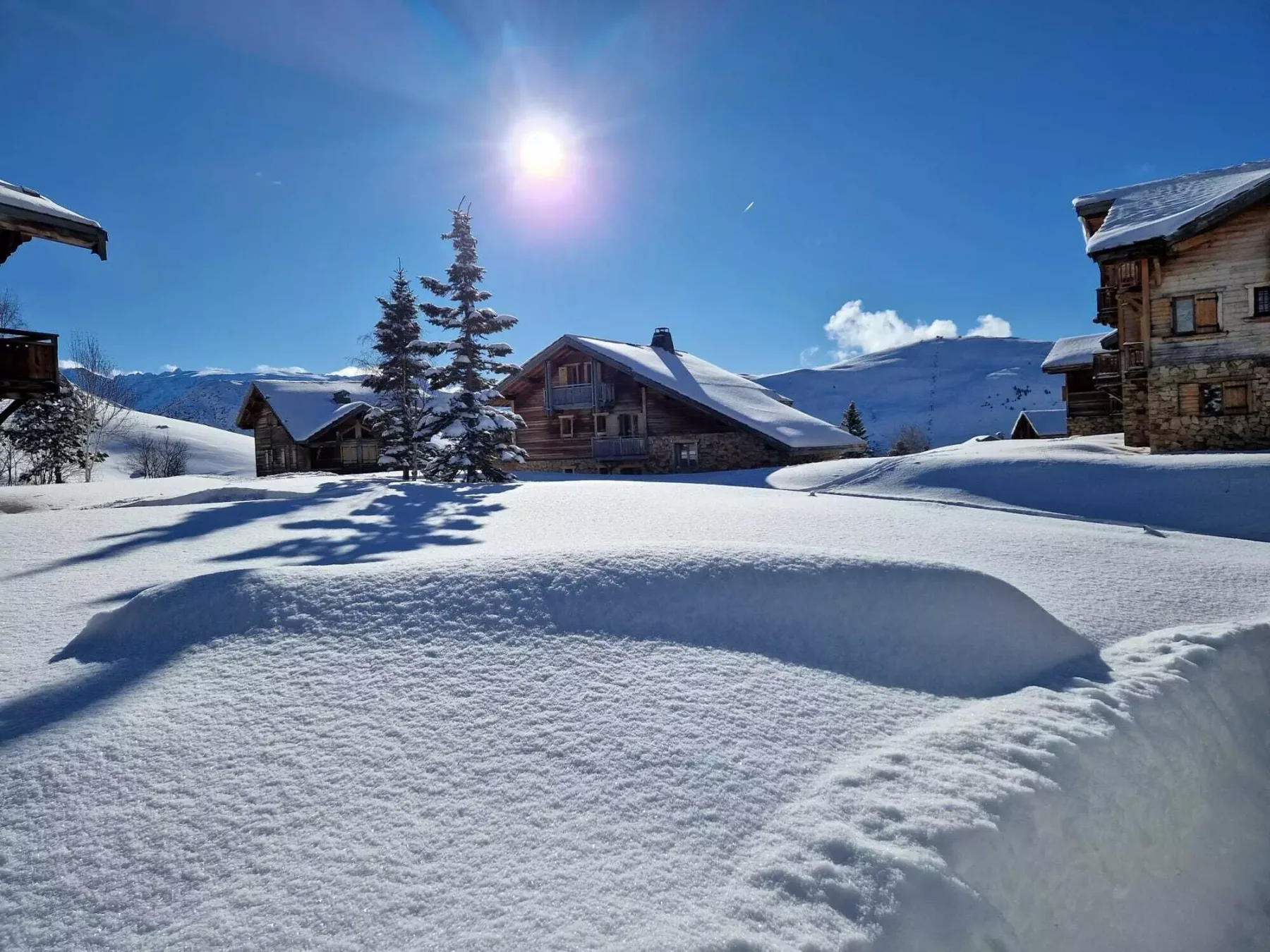 Snow-covered alpine chalets sit beneath a bright sun and mountain peaks.