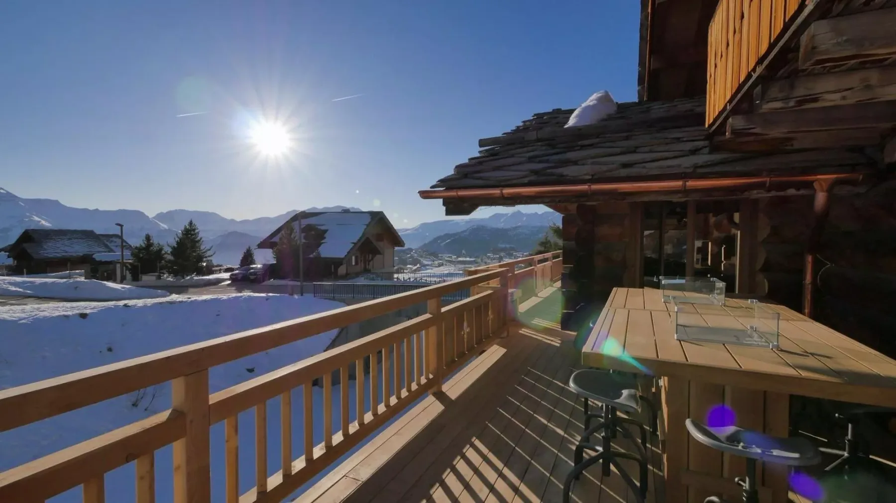 A wooden deck overlooks a snowy mountain landscape beneath a bright sun.