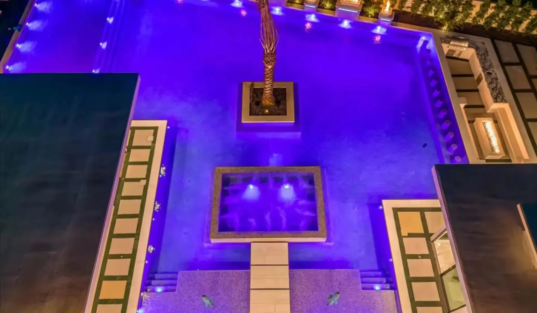 An illuminated swimming pool at night, with a small raised basin near the center.