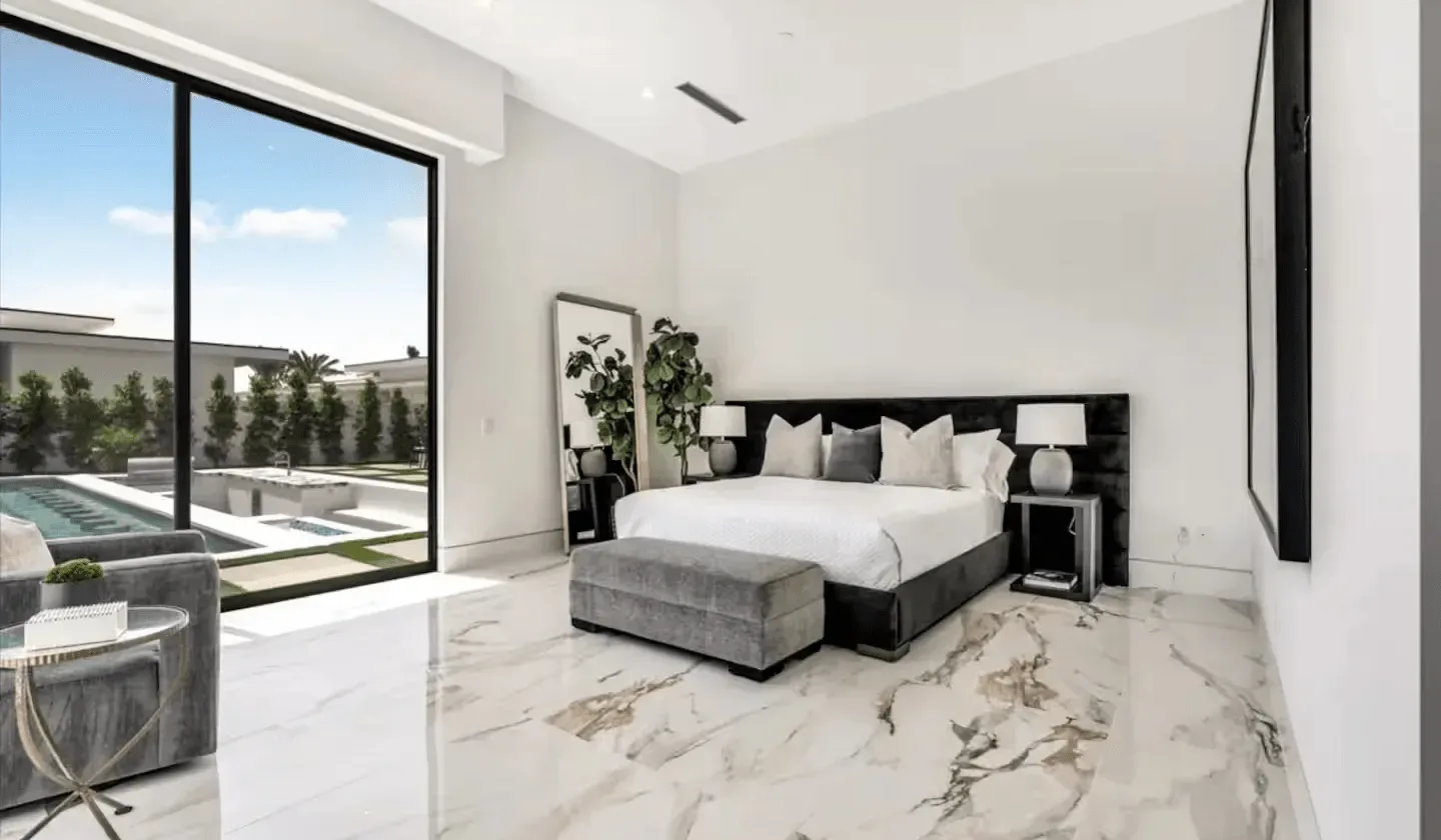 A bright bedroom has a large bed, gray seating, and floor-to-ceiling windows.
