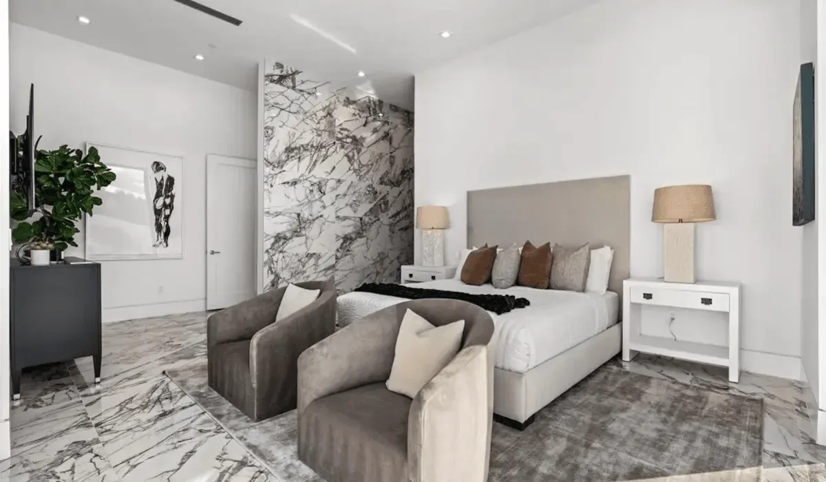 A bright bedroom with a bed, bedside tables, and two upholstered seats.