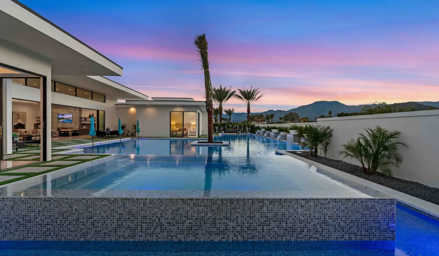 Una piscina junto a una casa moderna, con palmeras y montañas a lo lejos.