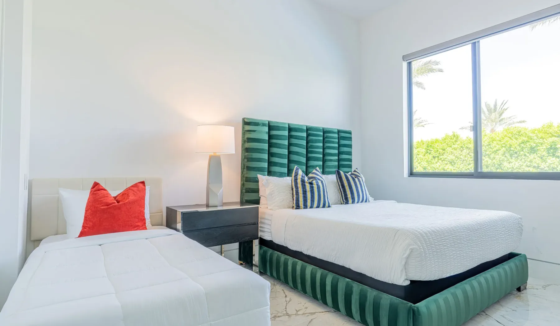 A bright bedroom with two beds, including a green upholstered bed beside a large window.