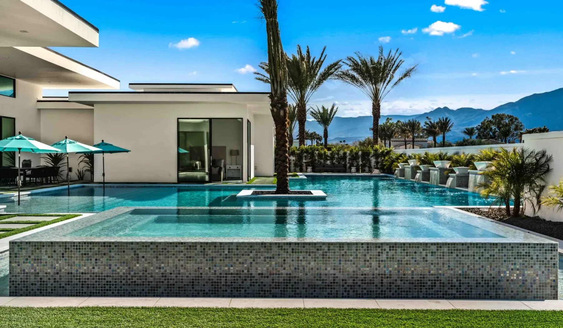 A modern outdoor swimming pool beside a white building, with palm trees and mountains beyond.