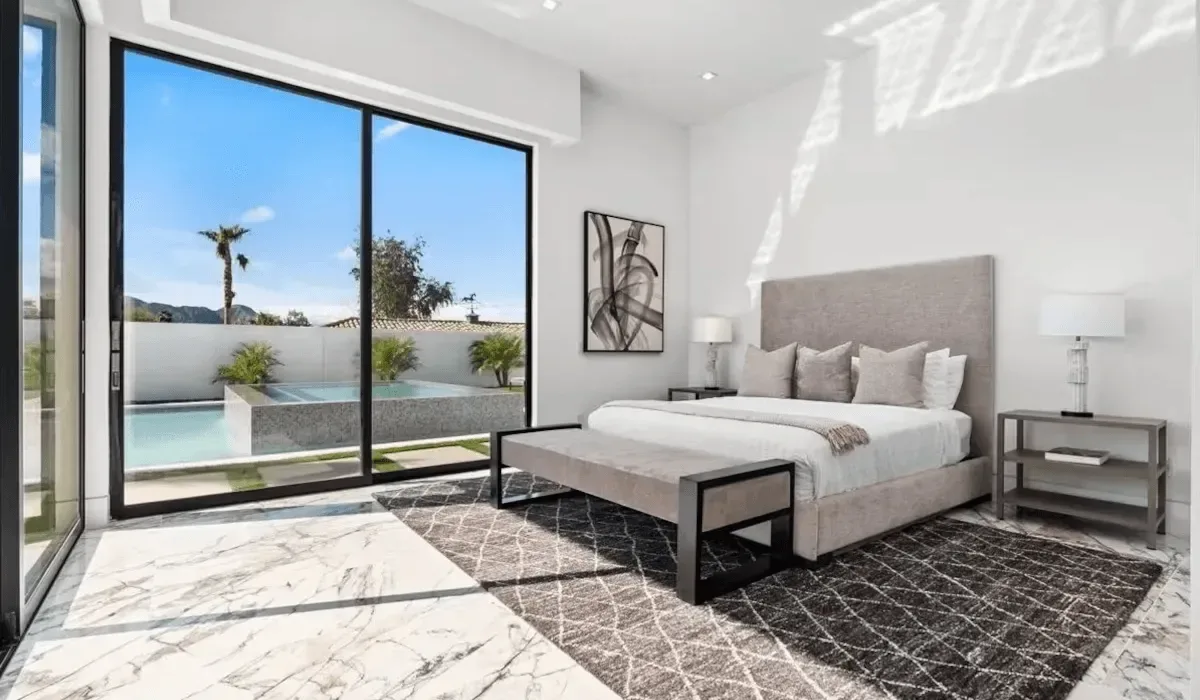 A bright bedroom with a large bed beside floor-to-ceiling windows overlooking a pool.