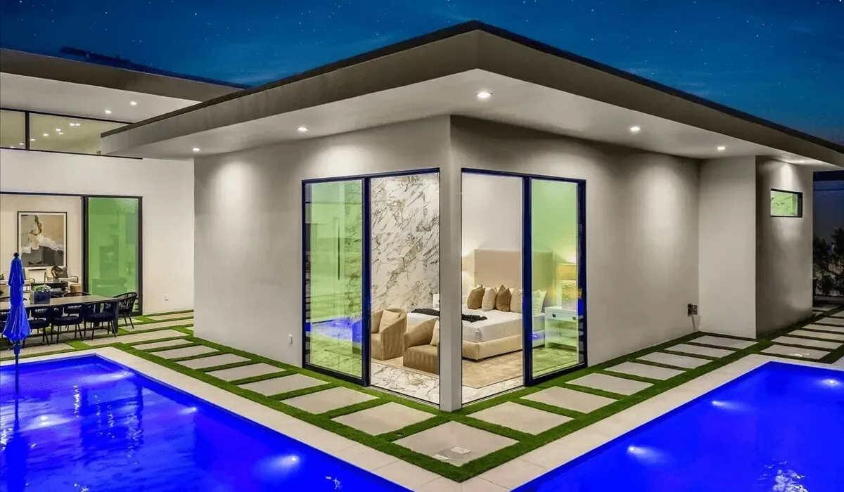 A modern house opens onto a swimming pool, with a bedroom visible through glass doors.