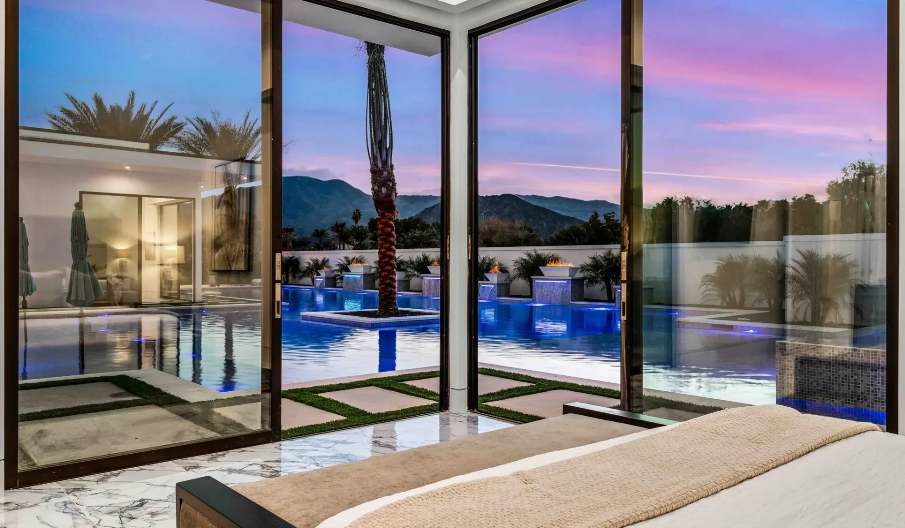 A bedroom opens through floor-to-ceiling windows onto a pool at sunset.