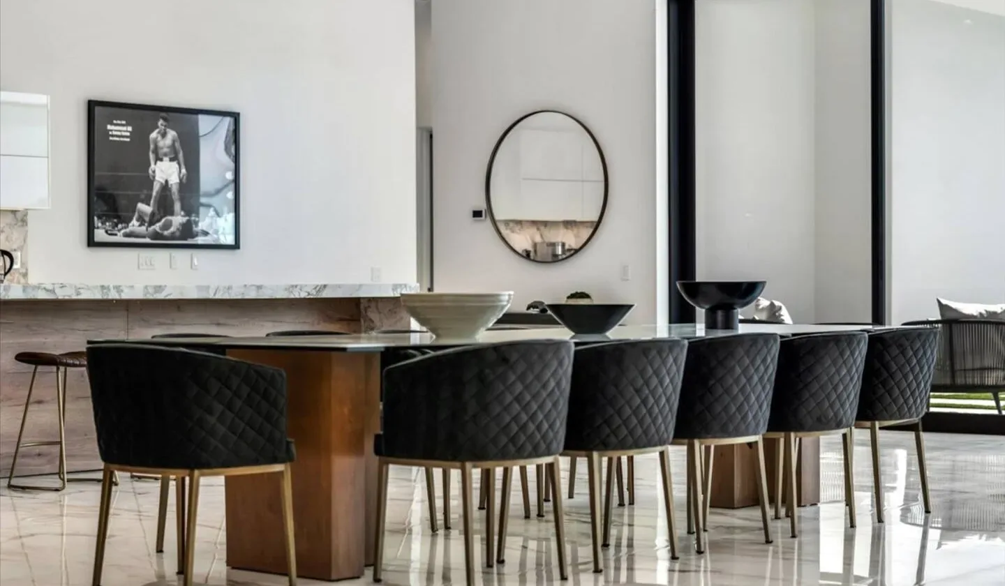A modern dining area features a long table with black seating and large windows.