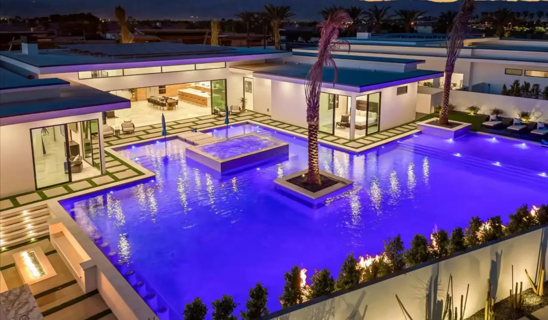 A modern villa surrounds a brightly illuminated swimming pool at dusk.