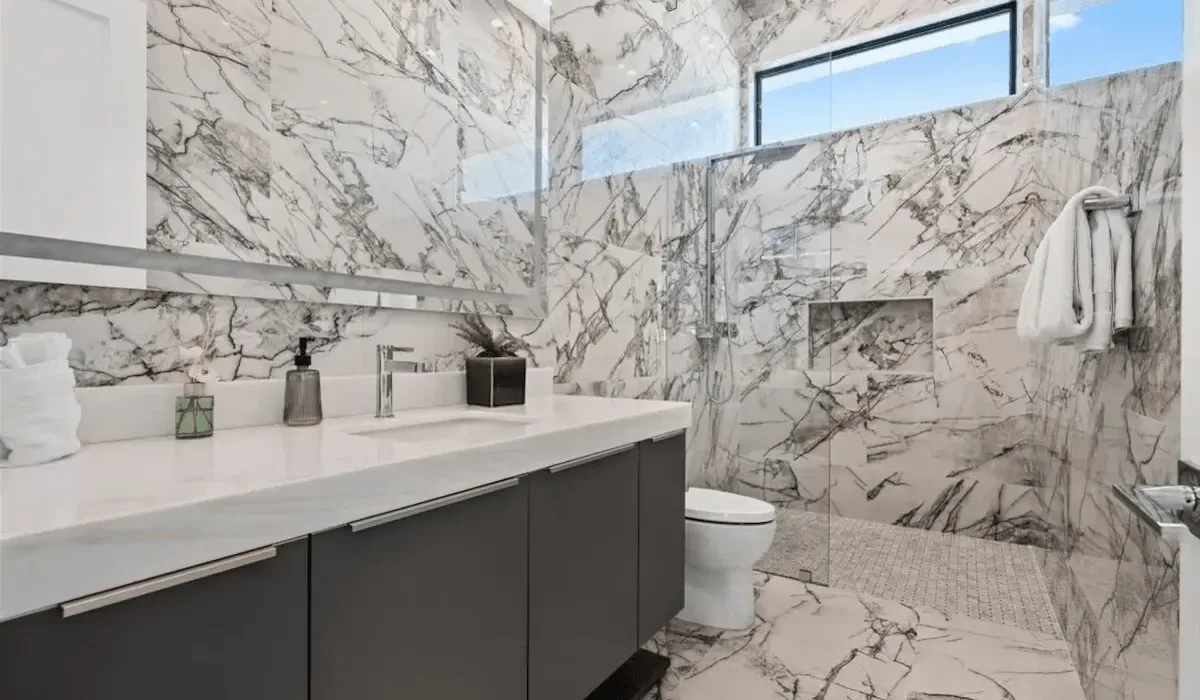 A marble-clad bathroom with a long vanity, toilet, and glass shower.