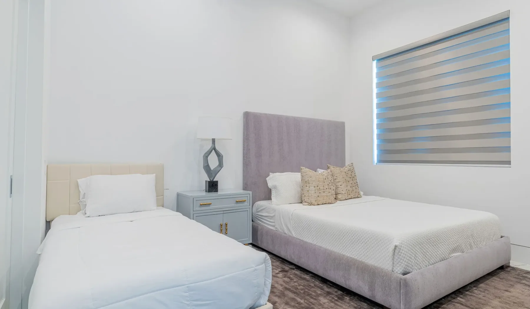 A bright bedroom with two beds and a window with striped blinds.