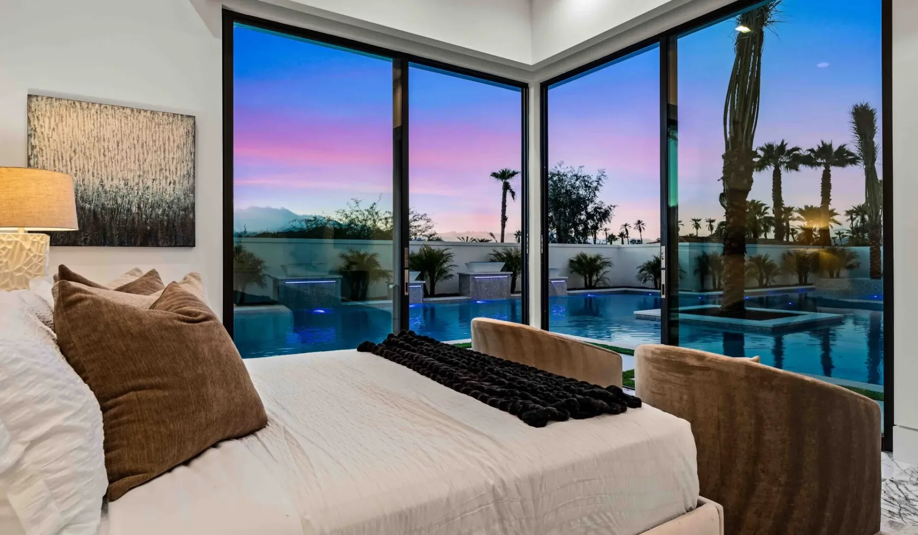 A bedroom with a bed beside large windows overlooking a pool at sunset.