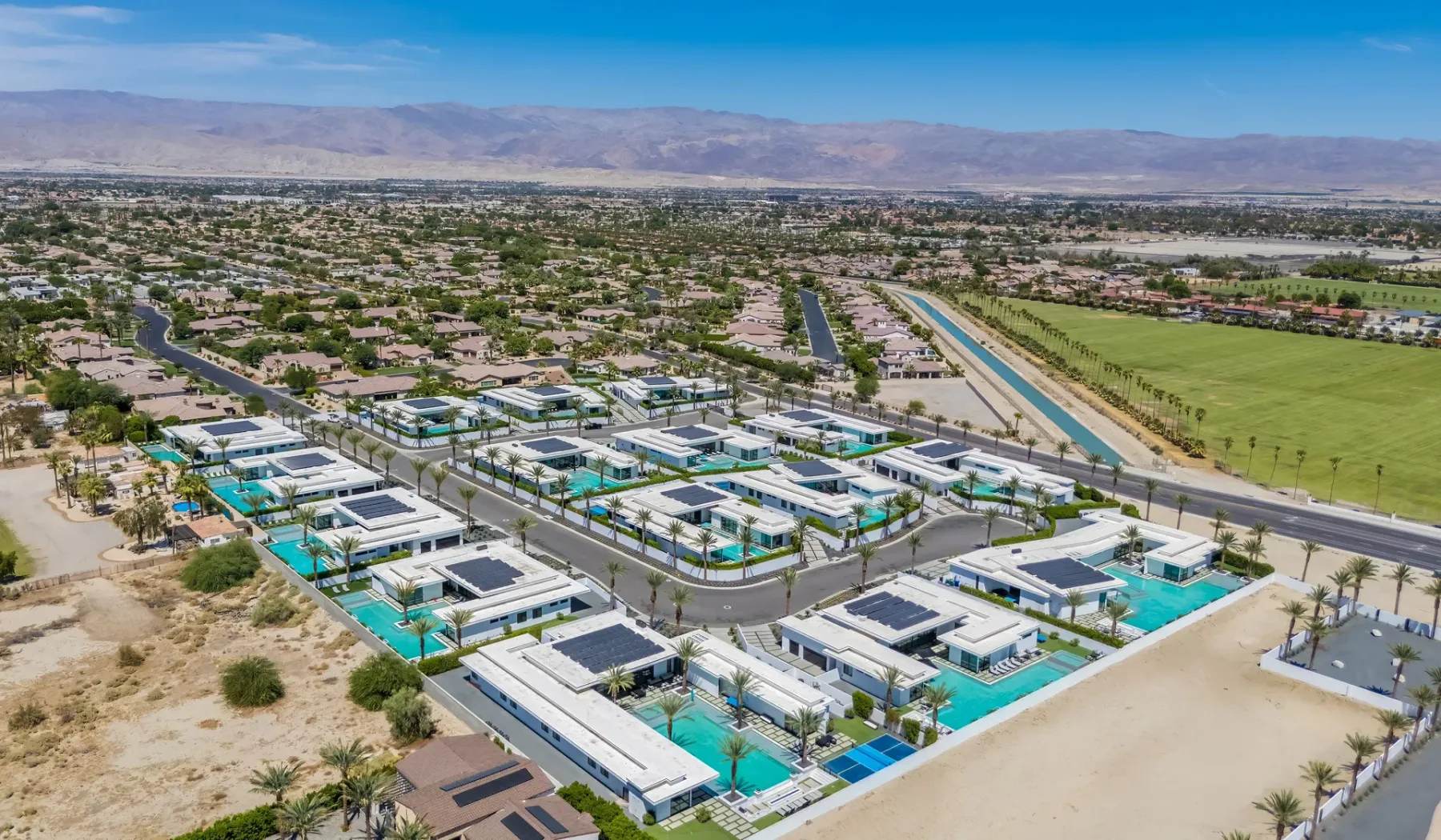 An aerial view shows a residential neighborhood with turquoise swimming pools and mountains beyond.