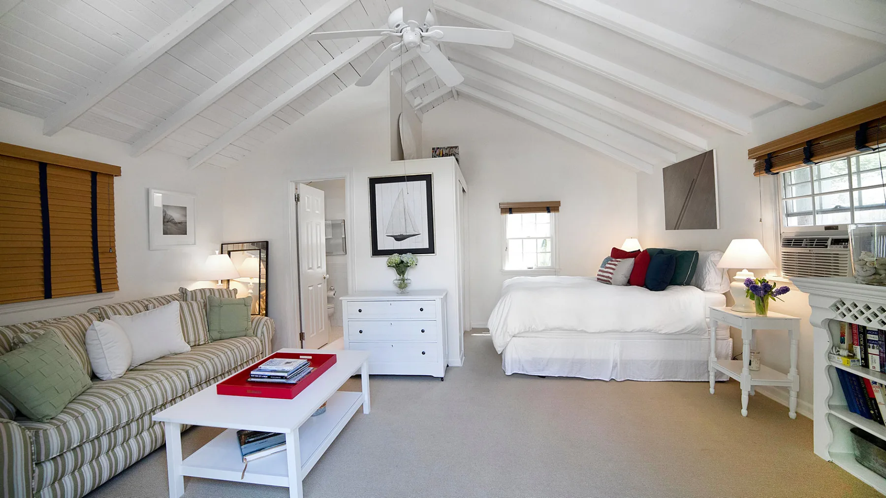 A bright bedroom with a bed, seating area, and white vaulted ceiling.