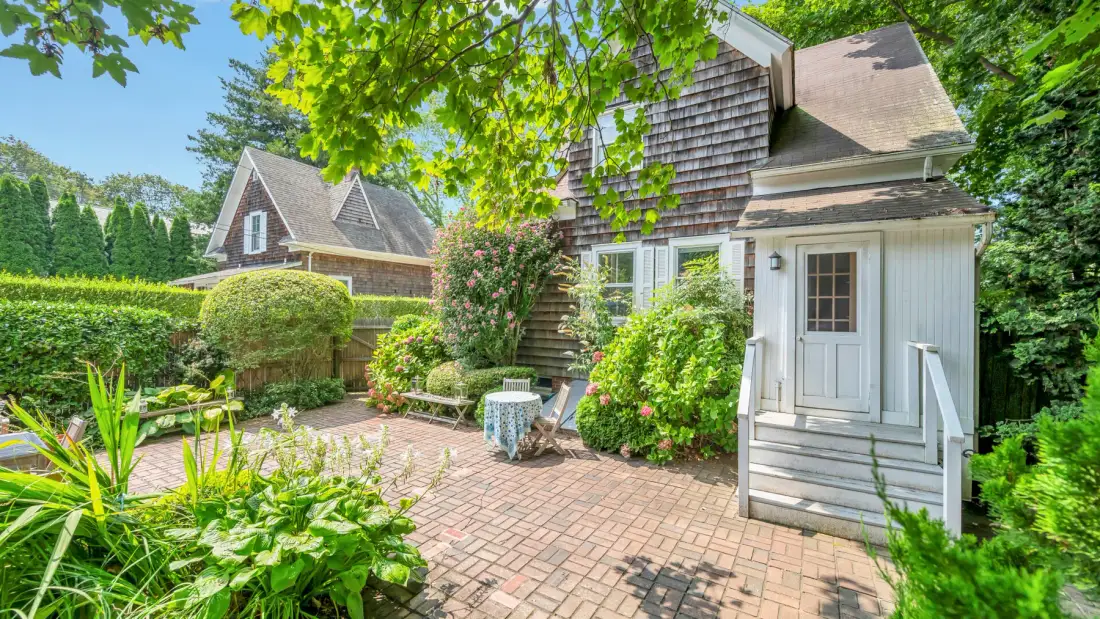 A landscaped brick patio sits beside a shingled cottage entrance, surrounded by greenery.