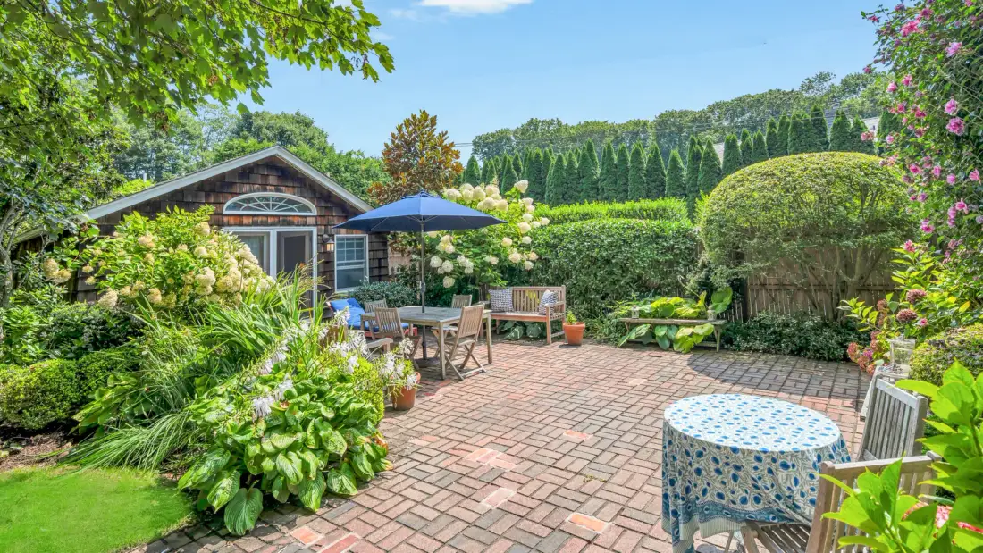 A brick patio garden with outdoor seating, an umbrella, and surrounding greenery.