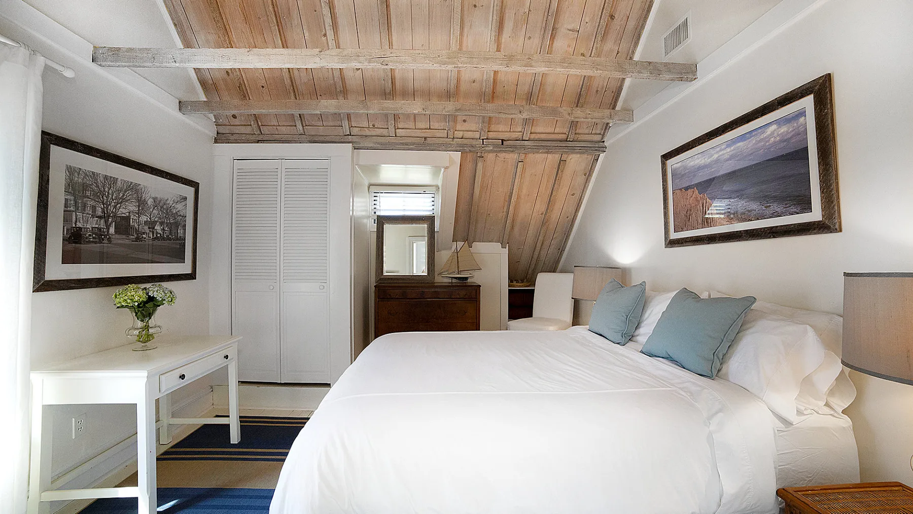 A bedroom with a white bed, blue pillows, and exposed wooden ceiling beams.