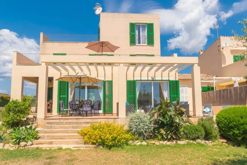 A cream-colored house with green shutters faces a garden and outdoor seating.