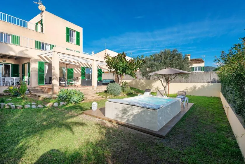 A sunny garden beside a modern house features a raised hot tub and patio seating.