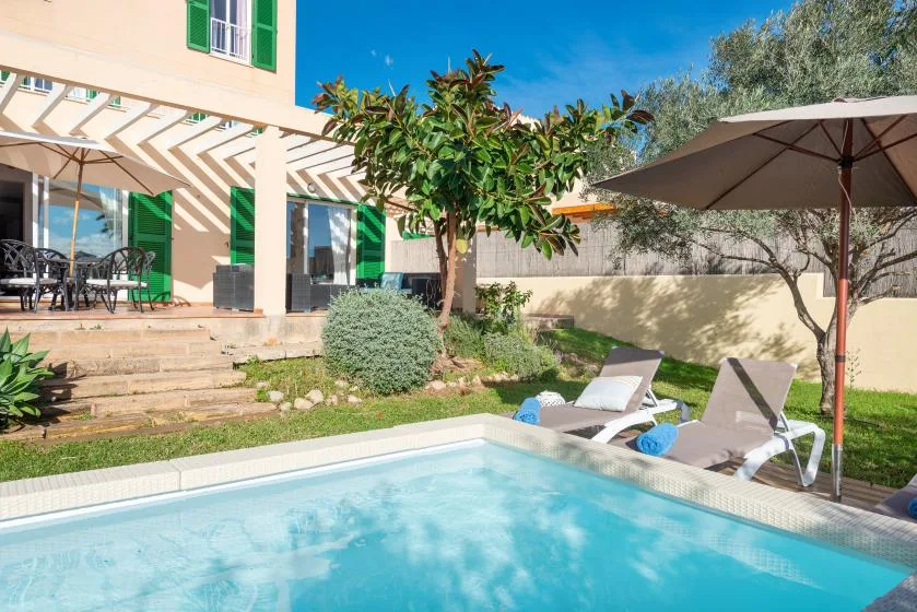 A sunny courtyard features a swimming pool beside a patio with seating and an umbrella.